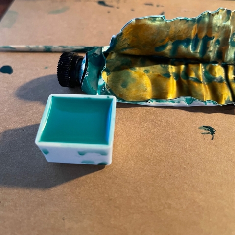 A half pan full of water and a little bit of green pigment, in front of a cut open tube of watercolour paint.